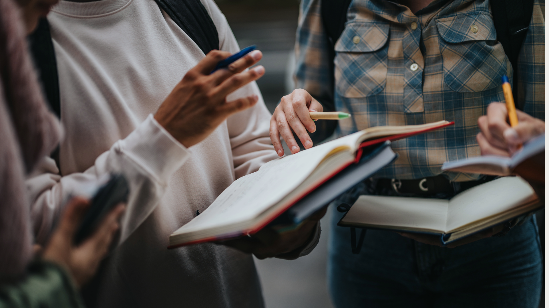 A close-up view of students engaged in a group discussion outdoors, using open books and notebooks. The scene conveys teamwork, learning, and academic collaboration.