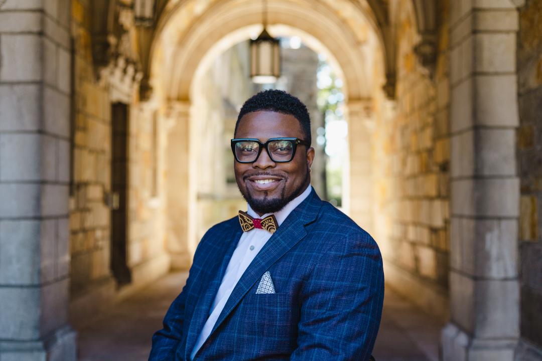 Person in a blue checkered suit with a bow tie standing under a stone archway.