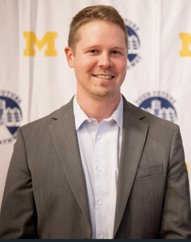 Smiling person standing in front of a white tarp that has the Michigan Block M on it. Person is wearing a collared dress shirt underneath a suit jacket.