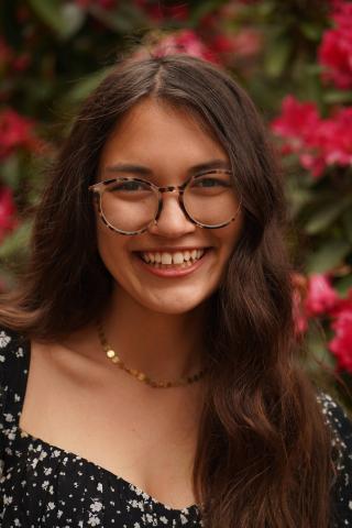 Close-up portrait of a young woman with long dark hair, wearing round tortoiseshell glasses, a black floral top, and a gold necklace, smiling with pink flowers in the blurred background.