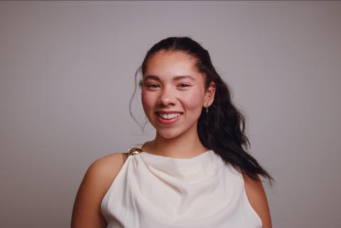 Portrait of a smiling person with dark hair in a ponytail, wearing a white draped sleeveless top with a gold shoulder clasp against a gray background.