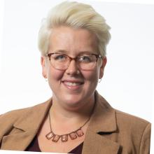 White woman with short blonde hair and glasses wearing a suit jacket and smiling on a white background