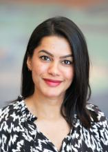 Female with dark hair wearing a black and white patterned shirt  standing in front of neutral background 