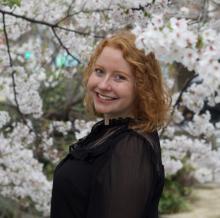 Person with red hair wearing a black shirt and smiling, with cherry blossoms in the background.