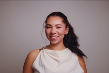 Portrait of a smiling person with dark hair in a ponytail, wearing a white draped sleeveless top with a gold shoulder clasp against a gray background.